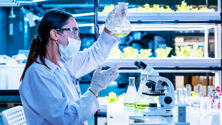 Científica con mascarilla usa pipeta en laboratorio con plantas y tubos de ensayo